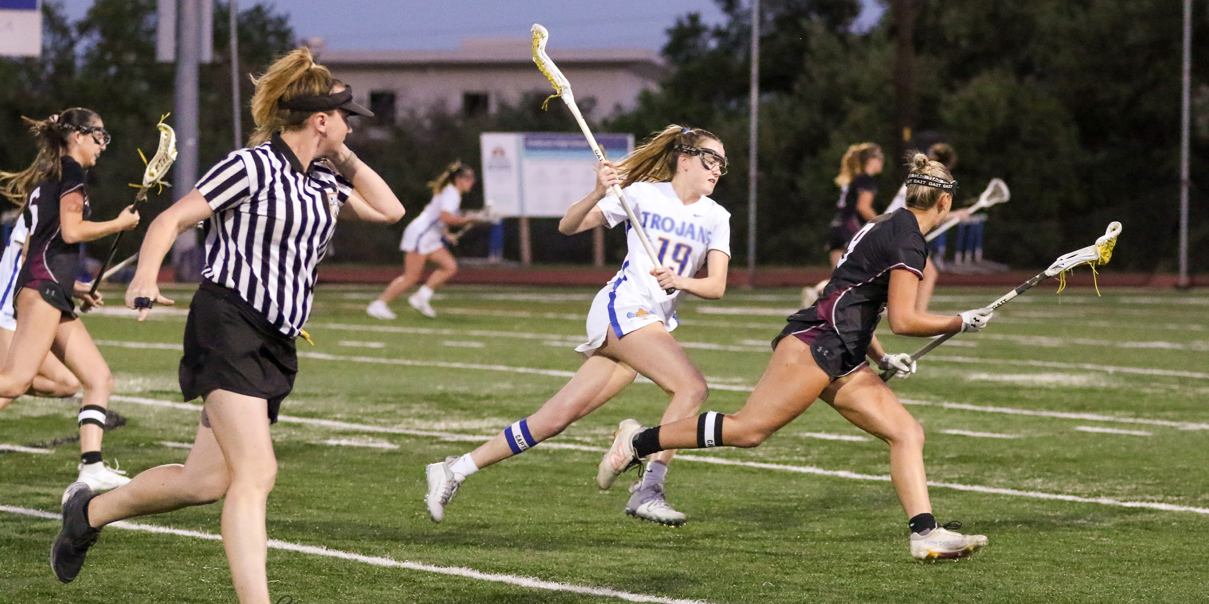 photo of lacrosse officials running while observing the interaction between two lacrosse players; one player has the ball and the other is trying to get in position to check the ball carrier's stick.